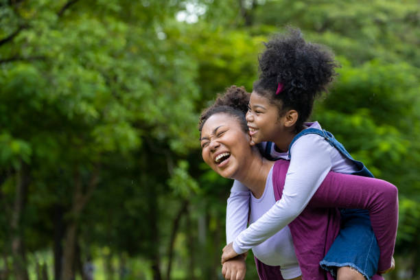 la madre afroamericana está jugando a montar en piggyback con su hija pequeña mientras hace un picnic de verano en el parque público para el concepto de bienestar y felicidad con espacio de copia - family single mother black mother fotografías e imágenes de stock