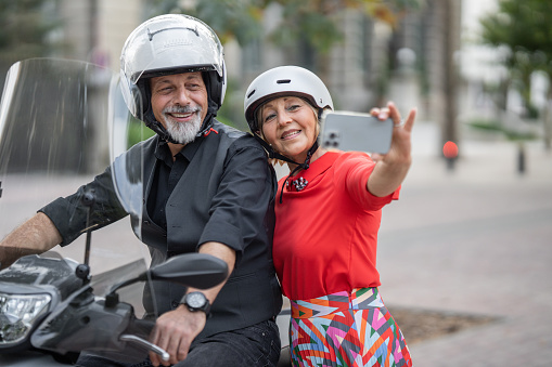 Mature couple with crash helmets preparing to go on a ride with motorcycle outdoors in city. Man is sitting on a bike, while woman is standing next to him and using phone to take a selfie.
