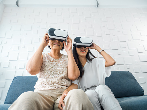 Asian young woman, happy daughter and her mother, old senior female wearing VR glasses, enjoy 3d game online together in white room. Mom and daughter with reality virtual technology lifestyle concept.
