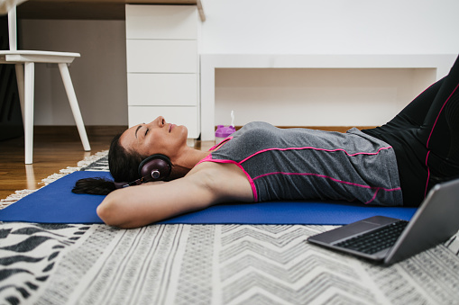Positive middle age woman lying on room floor and resting after successful home fitness workout. She listens to music with Bluetooth wireless headphones. Stay at home concept.