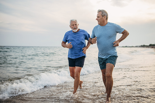 Two senior men jogging on the beach by the sea together, they are on vacation in Greece.