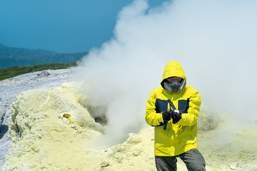 male volcanologist on the background of a smoking fumarole examines a sample of a sulfur mineral with a geological hammer