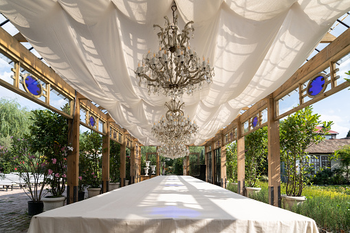 Decorated place prepared for wedding celebration in backyard near green trees growing in pots. Long table covered with white tablecloth stands under canopy and luxury chandeliers on sunny summer day