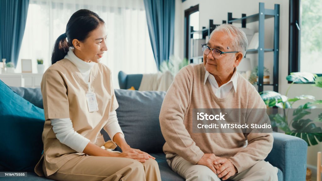 Young Asian Female professional caregiver take care older men share a story together in living room at home. Young Asian Female professional caregiver take care older men share a story together in living room at home. Girl personal care hold arm mature male patient in nursing house, Healthcare concept. Nursing Home Stock Photo