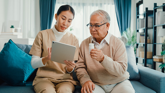 Young Asian female caregiver, personal care nurse teach older men use medicine indication and instruction application on digital tablet at home.