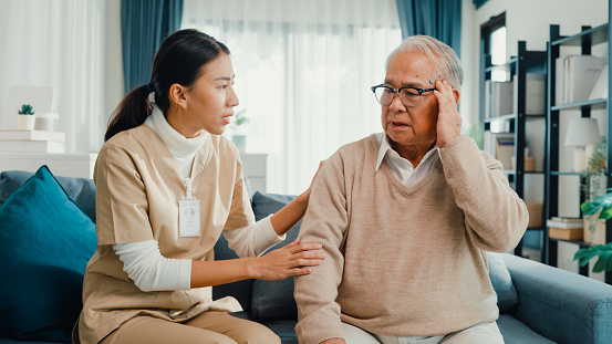 Young Asian female professional caregiver take care touch to calm sick elderly male patient in living room at home. Girl personal care support mature man patient in nursing house, Healthcare concept.