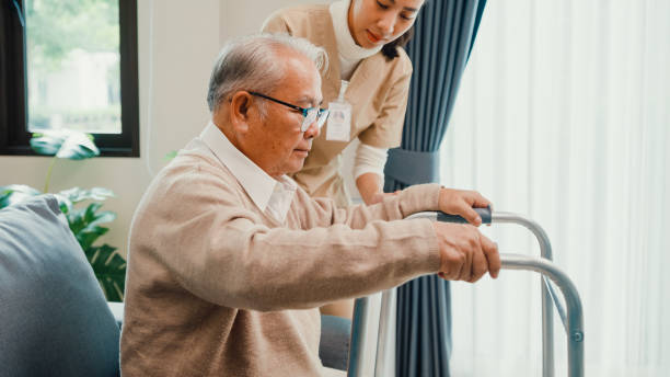 Young Asian female caregiver teach old male to walk use walker take care hold hand elderly patient in living room at home. Young Asian female caregiver teach old male to walk use walker take care hold hand elderly patient in living room at home. Assistance gentle girl support mature man, Rehabilitation healthcare concept. hip replacement stock pictures, royalty-free photos & images