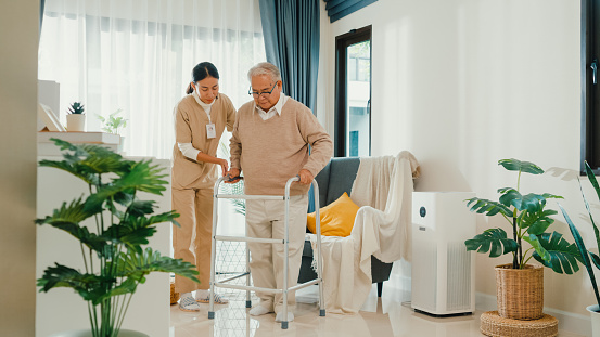Young Asian female caregiver teach old male to walk use walker take care hold hand elderly patient in living room at home. Assistance gentle girl support mature man, Rehabilitation healthcare concept.
