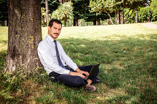 The young businessman is sitting on the lawn and leaning against a tree while he writes a work email.