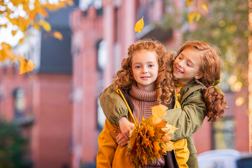 Two red-haired girls, sisters, cheerfully walk along city street during golden autumn leaf fall.
