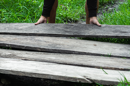 young latin guy laying some old gray wooden planks on a grassy land, construction of a wooden path or floor. concept of prosperity. scarce economic resources in a popular neighborhood