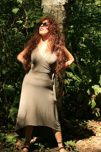 A Mexican woman against a tree in a forest with a ray of sunshine hitting her. She is wearing sunglasses and a an olive colored dress, with sandals.