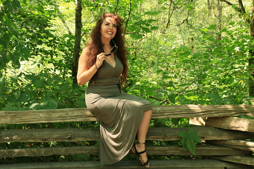 A Mexican model sitting on a cedar rail fence in a public park. She is wearing a long khaki green sleeveless dress, a necklace and sandals.