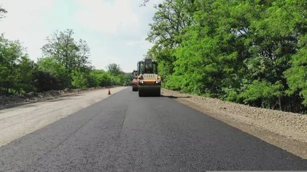 Photo of repair of a highway, roller compactor machine and asphalt finisher laying a new fresh asphalt pavement, covering on one side of the traffic. Road construction works