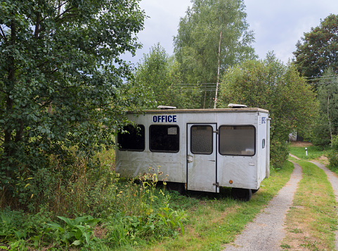 old small white trailer house with blue sign office, abandoned caravan in rural landscape near footpath, green grass and trees, blue sky