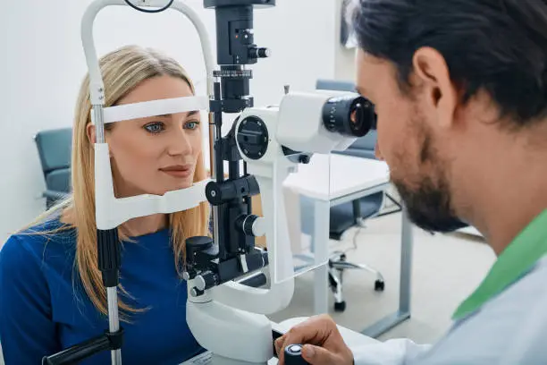Beautiful woman while eye exam with binocular slit-lamp at ophthalmology clinic with experienced male optometrist