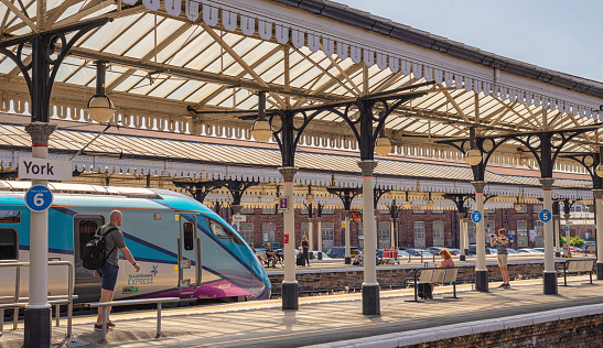 York, UK.  August 11, 2022.  A train rests under an old 19th Century historic canopy. Passengers are on the platform and a youth takes a photograph of the locomotive.