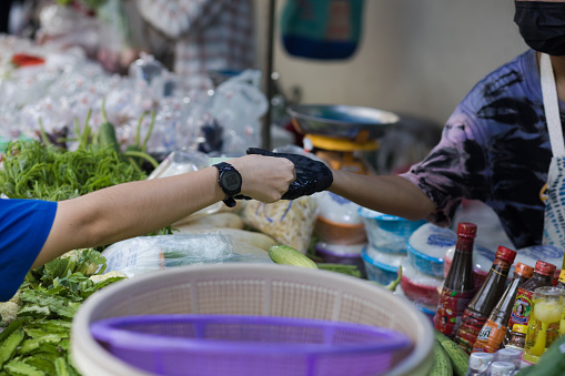 Thai woman is paying cash to vendor woman at food market stall. Vendor woman is wearing a glove at right hand. Scene is on local market residential district of Bangkok Chatuchak