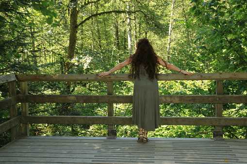 A Mexican model looking out onto a public park that is still forest. She is wearing a long sleeveless khaki green dress and sandals.