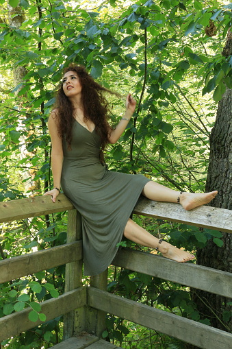A Mexican woman sitting on the top rail of a viewpoint in a public park. She is wearing a sleeveless, long, olive green dress and is barefoot. She also has ankle bracelets.