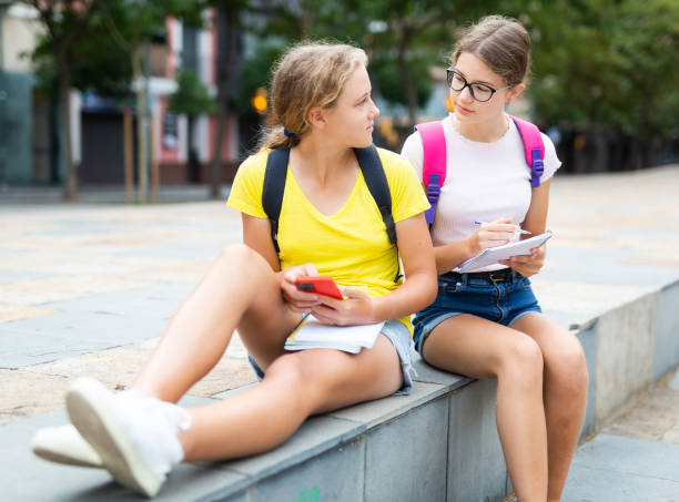 duas alunas usam telefones para verificar aulas e escrever tarefas completas em caderno - 12 15 months - fotografias e filmes do acervo