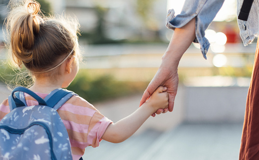 Back view of little girl with backpack holding a mom hand while walking to the school together.