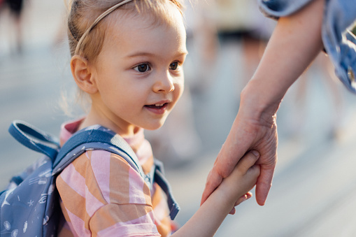 Close up photo of little girl with backpack holding a mom hand while walking to the school together.