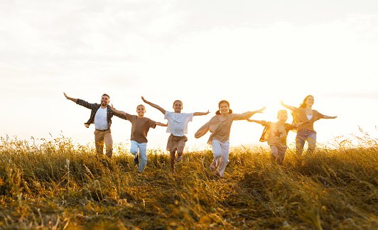 Happy young parents with children in casual clothes running with outstretched arms while enjoying time together on meadow in summer evening