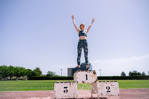 Low angle of female athlete in sportswear raising arms while standing on rusty first place podium after victory in track and field competition on summer day on stadium