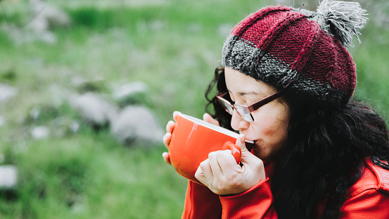 Latin brunette woman, wearing a red leather jacket, glasses and a wool cap, sipping hot coffee in nature.