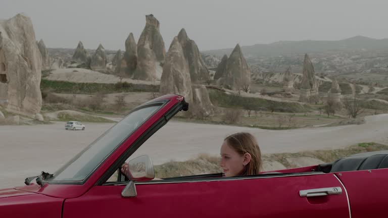 Young smiling girl sitting in red retro cabriolet car in valley of Cappadocia, looking around and enjoying beautiful scenery. Side view.