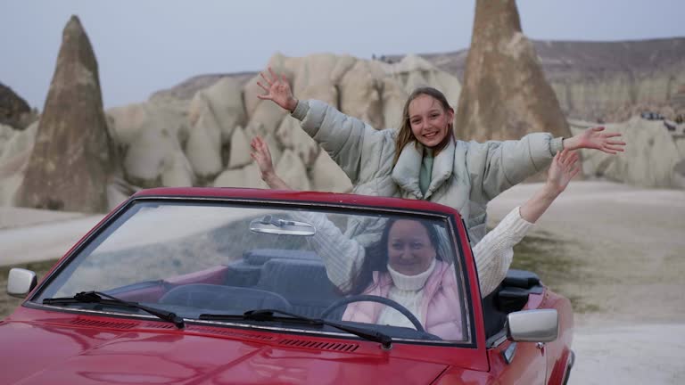 Happy woman and playful girl swaying hands in red retro cabrio car against rock formations background in stone valley Cappadocia in Turkey. Family travel concept.