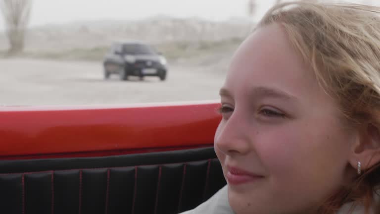 Close-up face of teenage girl with hair fluttering in wind in moving red convertible car among sand hills and dry bushes enjoying scenery. Road trip concept.