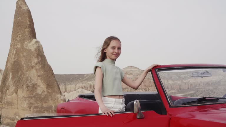 Pretty young girl stand by open door of red retro cabrio car against rock formations, mountains and grey sky in Cappadocia Turkey. Wind in hair. Travel and car rent concept.