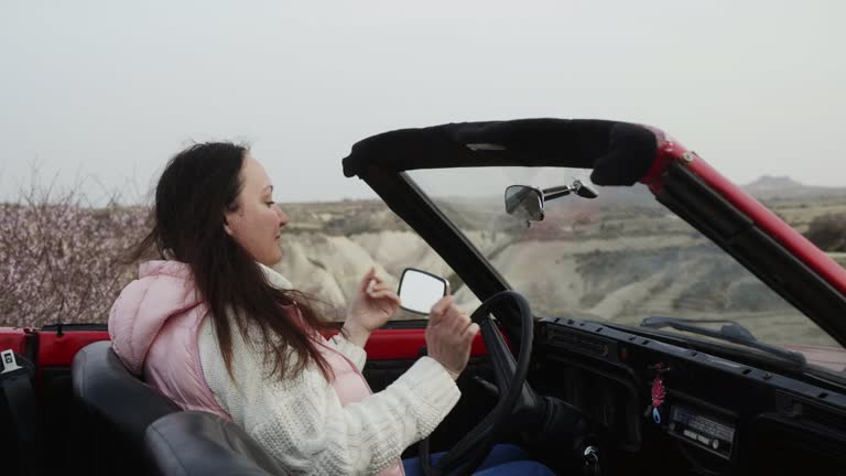 Woman is Sitting in her Red Retro Cabriolet Looking in Mirror Doing her Hair.