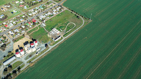 Expansive Aerial View Of Rolling Green Agricultural Fields, Farm Buildings, And A Long Country Road Under A Cloudy Sky.