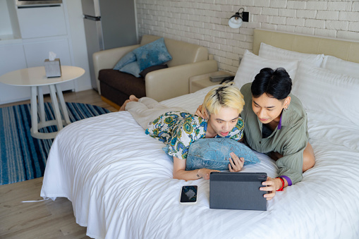 Asian Gay couple happily using tablet for entertainment on vacation in hotels room.