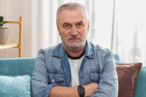 hombre mayor enojado con cabello gris y brazos cruzados mirando a la cámara, sentado en el sofá de su casa - sulking fotografías e imágenes de stock