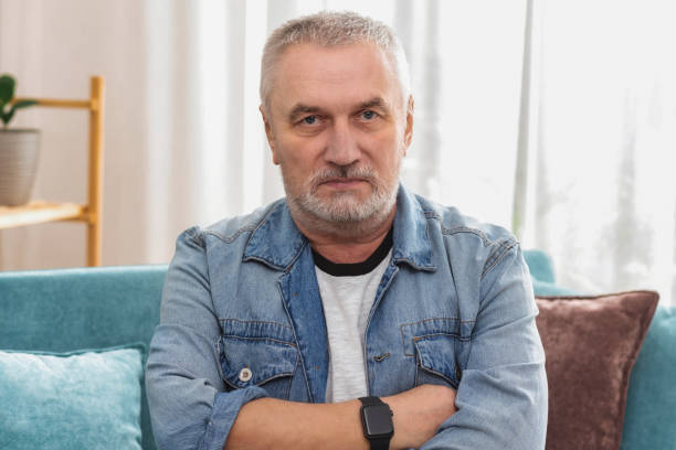 uomo anziano arrabbiato con i capelli grigi e le braccia incrociate che guarda la telecamera, seduto sul divano di casa - sullen foto e immagini stock