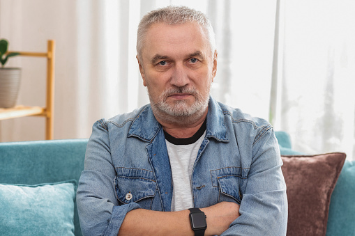 Angry senior man with gray hair and crossed arms looking at camera, sitting on sofa at home