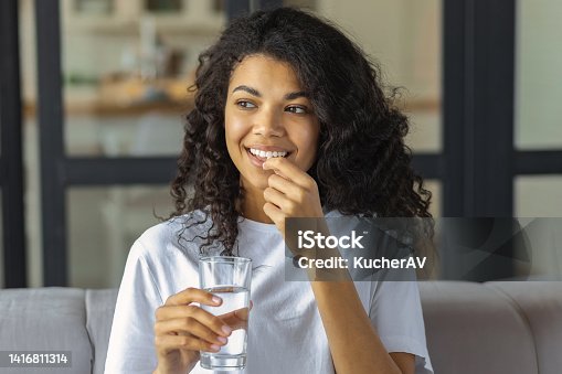 istock Young smiling African American woman holding vitamin pill and glass of water. Healthy lifestyle or diet nutrition concept 1416811314