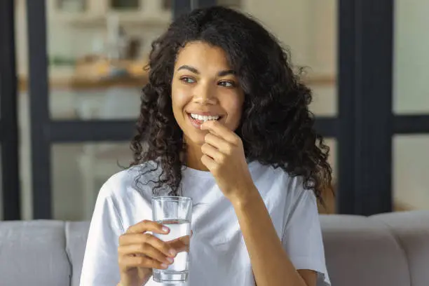 Young smiling African American woman holding vitamin pill and glass of water. Healthy lifestyle or diet nutrition concept