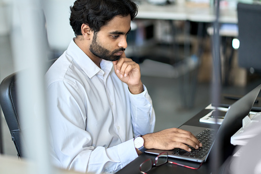 Busy young indian business man employee working using laptop, analyzing financial market, developing online project data, thinking on plan looking at computer, doing accounting management in office.