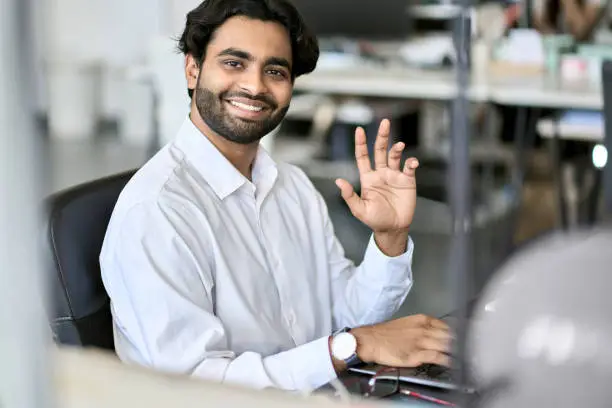 Happy indian young business man professional manager, office employee worker, programmer or analyst working online on laptop computer looking at camera waving hand sitting at workplace, portrait.