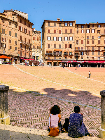 Siena, Tuscany, Italy, July 07 -- A couple of young women admire Piazza del Campo sitting on ground, in the historic heart of the medieval city of Siena, in Tuscany. Center of the ancient medieval city since 1169, the Piazza del Campo or simply the Campo is one of the most beautiful and famous squares in the world for its particular shell shape. In this space every year the seventeen historical villages of Siena compete in the Palio, one of the oldest horse races in the world. Siena is one of the most beautiful Italian cities of art, in the heart of the Tuscan hills, visited for its immense artistic and historical heritage and for its famous popular traditions. Since 1995 the historic center of Siena has been declared a World Heritage Site by UNESCO. Image in high definition format.