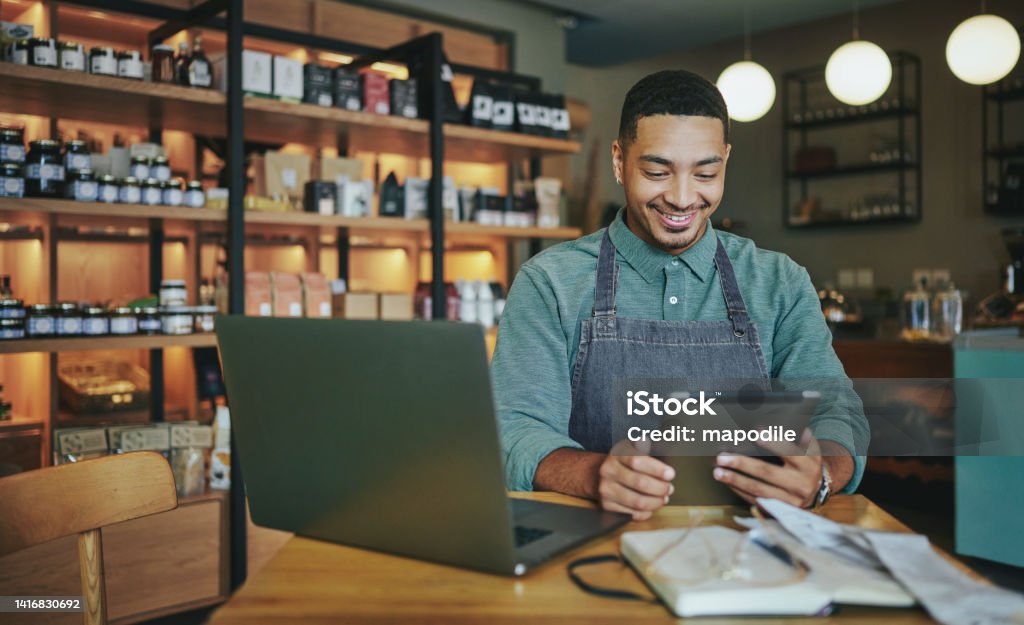 Smiling deli manager working on a tablet and laptop in his shop Smiling young male deli owner working a tablet and a laptop while sitting at a table in his artisanal shop Small Business Stock Photo