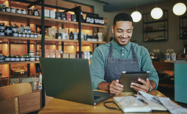 Smiling deli manager working on a tablet and laptop in his shop Smiling young male deli owner working a tablet and a laptop while sitting at a table in his artisanal shop small business stock pictures, royalty-free photos & images
