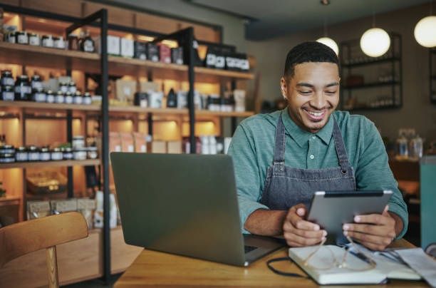 Deli manager smiling while working on a tablet and laptop in his store stock photo