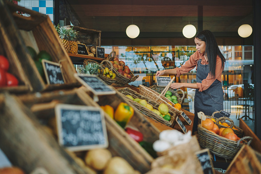 Young female supermarket owner organizing signs for organic fruit and vegetables and fruit on display in baskets outside her shop