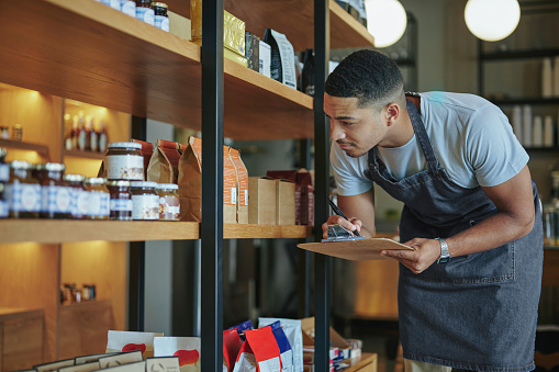 Young male delicatessen owner holding a clipboard taking inventory of artisanal merchandise on shelves in his shop